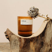 A brown soy wax candle with a label reading 'judaean', displayed on a wooden slice alongside a dried flower decoration.