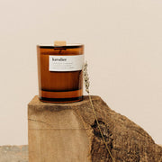 A medium amber soy candle with a wooden lid, sitting on a rustic wooden surface, with a white label that reads 'kavalier'.