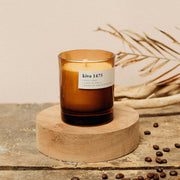 A medium amber colored candle with a label reading 'kiva 1475', placed on a wooden platform with coffee beans scattered around, against a neutral background.