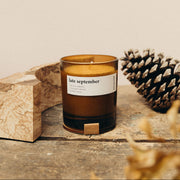 A soy candle with a label reading 'late september' emits smoke, placed on a wooden surface with a pine cone and piece of driftwood nearby.