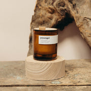 A brown amber soy candle in a clear glass jar with a wooden lid, labeled 'passenger', displayed on a wooden platform with a piece of driftwood in the background.