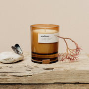 A bronze-colored soy candle with a label reading 'seafarer', placed on a wooden surface with a small white shell to the left and dried plant foliage to the right.